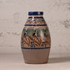 A handmade ceramic vase with a natural clay body, decorated with traditional hand-painted floral motifs in blue, green, and ochre orange, set against a rustic textured plaster wall.