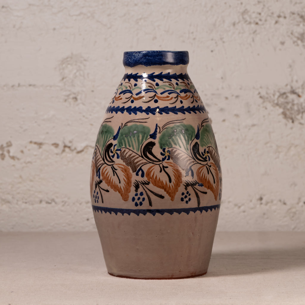 A handmade ceramic vase with a natural clay body, decorated with traditional hand-painted floral motifs in blue, green, and ochre orange, set against a rustic textured plaster wall.