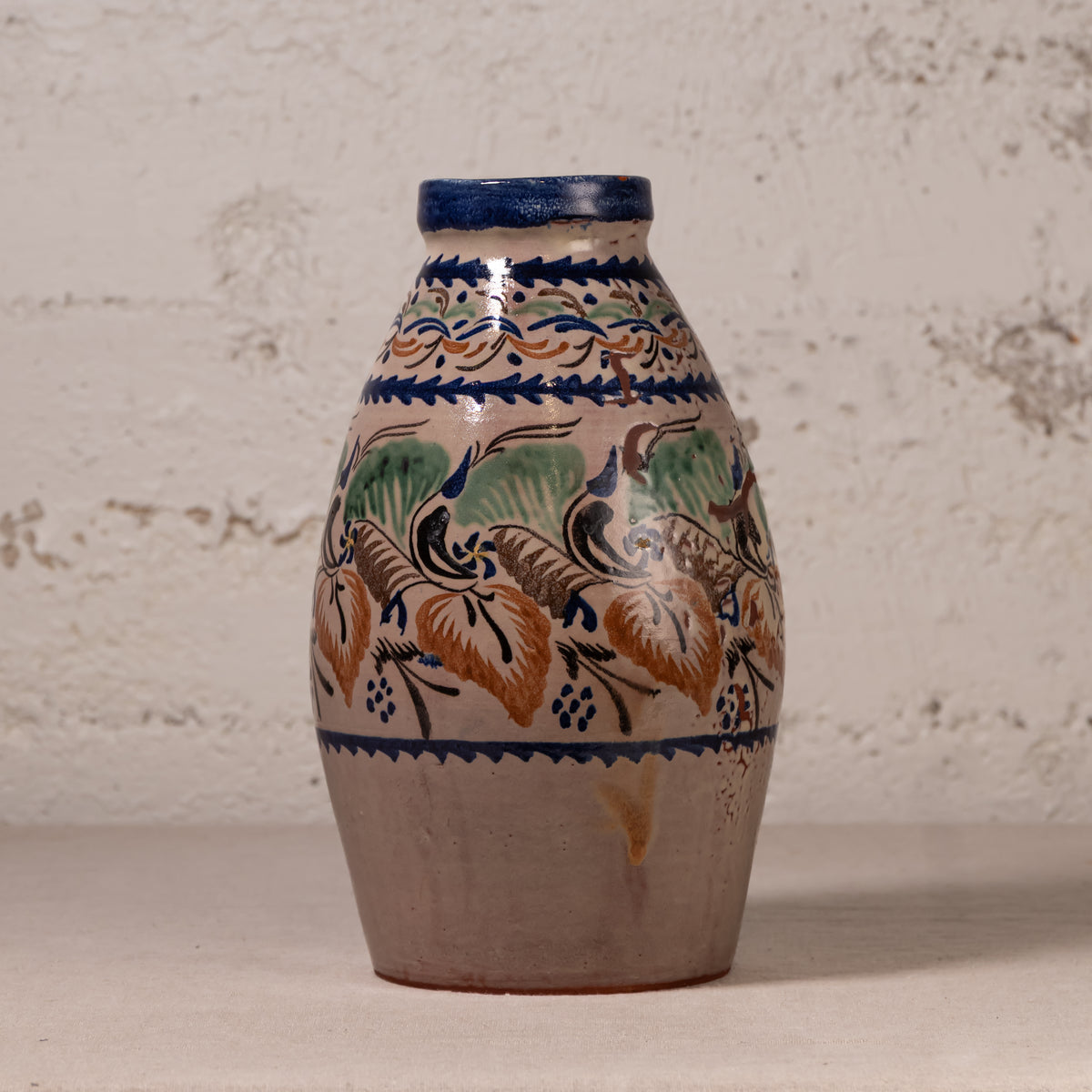 A handmade ceramic vase with a rustic, hand-painted floral pattern in blue, green, and ochre brown on a beige background, set against a textured off-white wall.