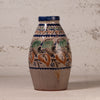 A handmade ceramic vase with a rustic, hand-painted floral pattern in blue, green, and ochre brown on a beige background, set against a textured off-white wall.