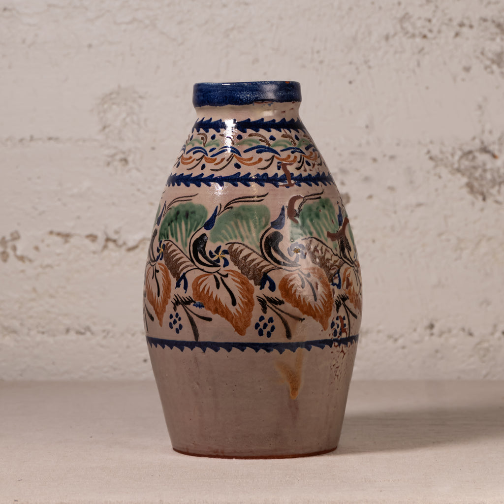 A handmade ceramic vase with a rustic, hand-painted floral pattern in blue, green, and ochre brown on a beige background, set against a textured off-white wall.