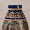Close-up of a hand-painted ceramic vase or bottle with a cream-colored glaze, decorated with blue, green, and orange traditional patterns, set against a neutral textured background.