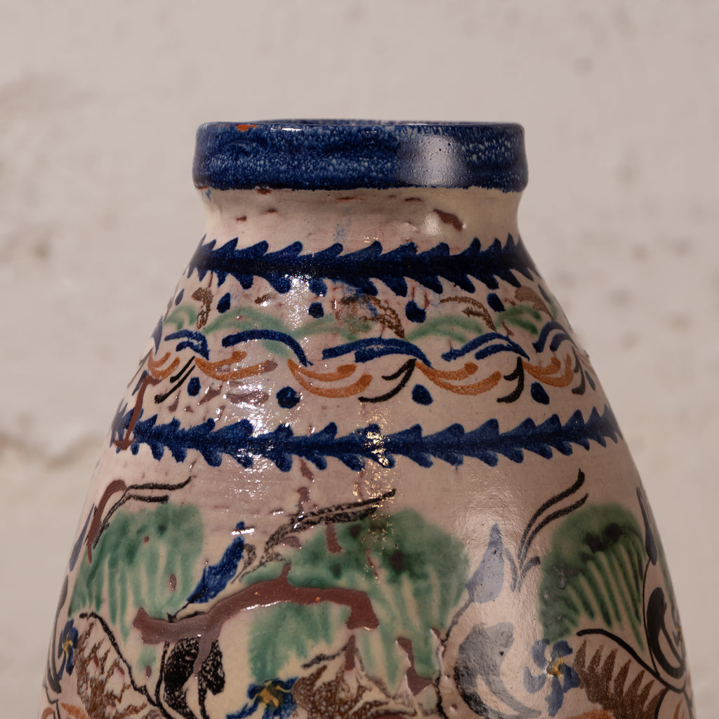 Close-up of a hand-painted ceramic vase or bottle with a cream-colored glaze, decorated with blue, green, and orange traditional patterns, set against a neutral textured background.