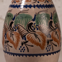 Close-up of a ceramic vase or jug with a hand-painted Mediterranean style pattern featuring blue birds, green leaves, and orange-brown grapes on a cream background.