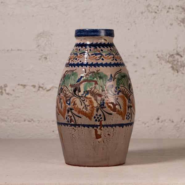 A vintage ceramic vase with a bulbous shape, featuring a hand-painted floral and bird design in blue, green, and brown on a beige stoneware base, set against a textured off-white wall.