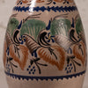 Close-up of a ceramic vase or jug with a hand-painted Mediterranean style pattern featuring blue birds, green leaves, and orange-brown grapes on a cream background.