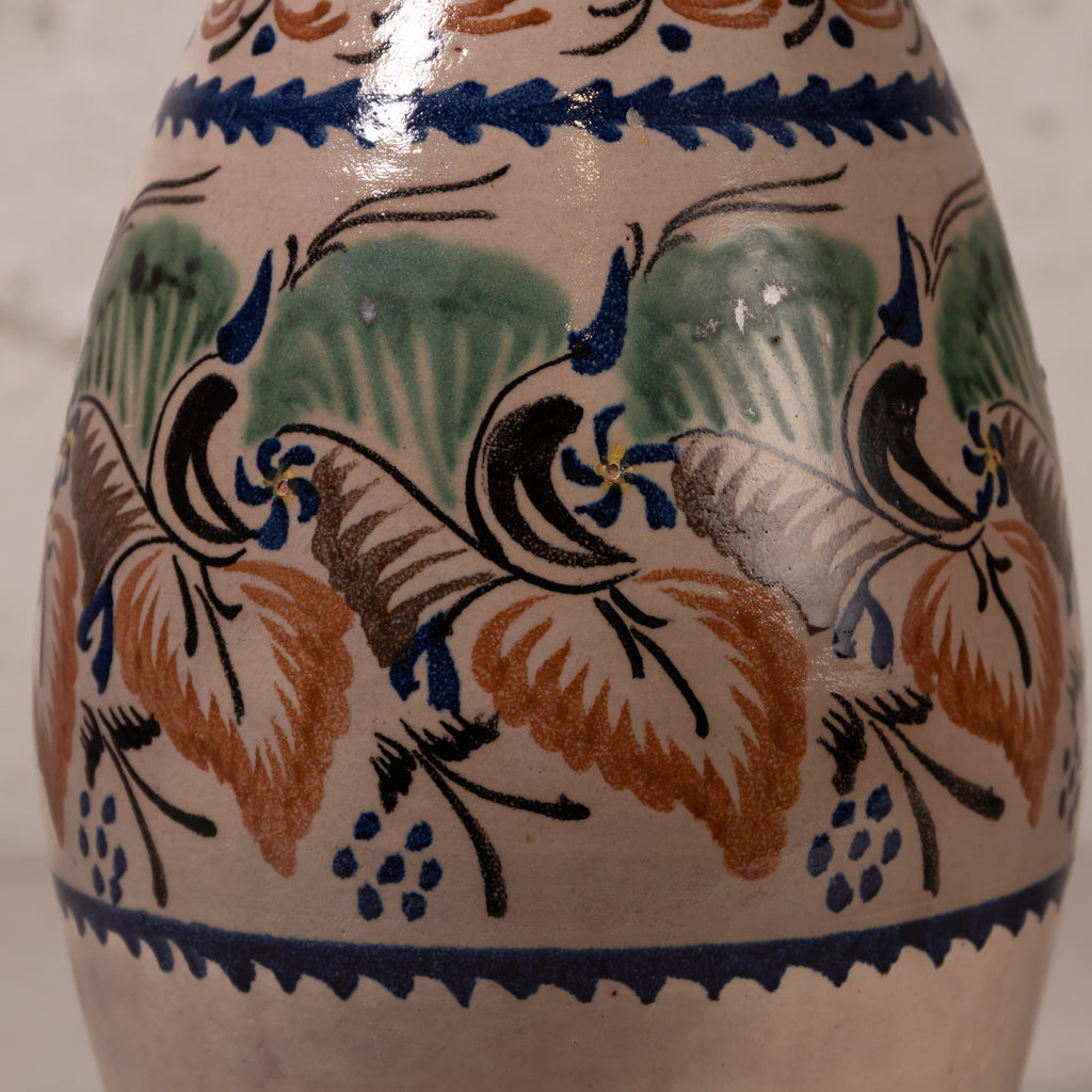 Close-up of a ceramic vase or jug with a hand-painted Mediterranean style pattern featuring blue birds, green leaves, and orange-brown grapes on a cream background.