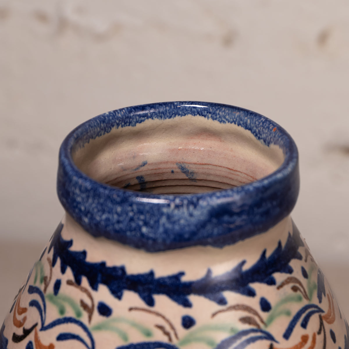Close-up of a ceramic vase or pot showing the glazed rim and upper body, decorated with a cream base and blue, green, and brown floral scroll patterns.