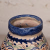 Close-up of a ceramic vase or pot showing the glazed rim and upper body, decorated with a cream base and blue, green, and brown floral scroll patterns.