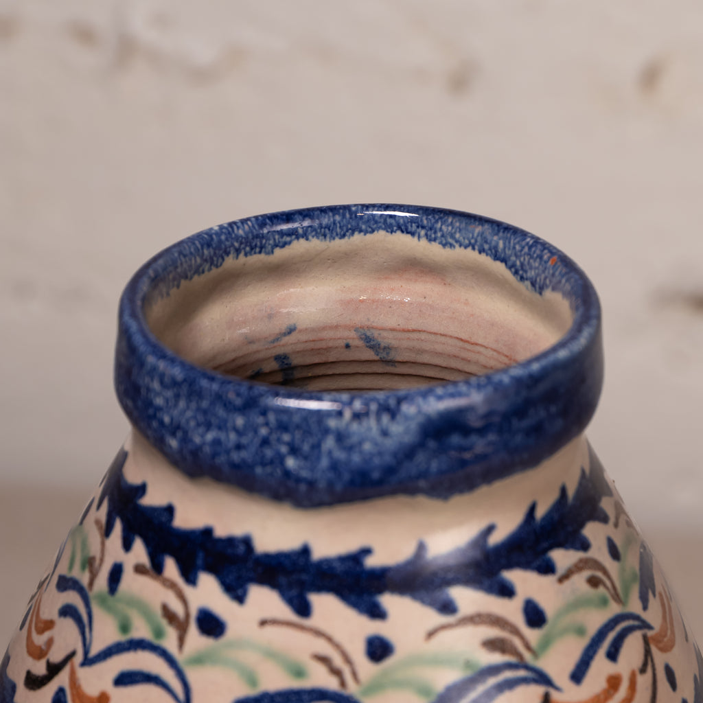 Close-up of a ceramic vase or pot showing the glazed rim and upper body, decorated with a cream base and blue, green, and brown floral scroll patterns.