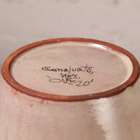 Close-up of the bottom of a handmade ceramic pot, showing a light pinkish-beige glazed interior with a terracotta rim and handwritten black ink signature reading 'Guajuala, MEX. CAPELO' on a neutral beige background.