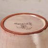 Close-up of the bottom of a handmade ceramic pot, showing a light pinkish-beige glazed interior with a terracotta rim and handwritten black ink signature reading 'Guajuala, MEX. CAPELO' on a neutral beige background.