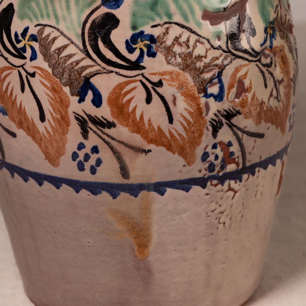 Close-up of a ceramic vase or pot with a hand-painted floral pattern in blue, brown, and green on a warm off-white glaze.
