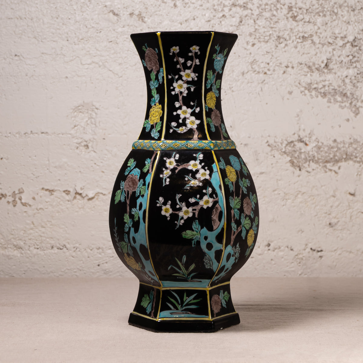 A black ceramic vase with a flared neck and hexagonal base, decorated with hand-painted floral motifs in turquoise, gold, green, and white, set against a textured off-white wall.