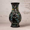 A black ceramic vase with a flared neck and hexagonal base, decorated with hand-painted floral motifs in turquoise, gold, green, and white, set against a textured off-white wall.