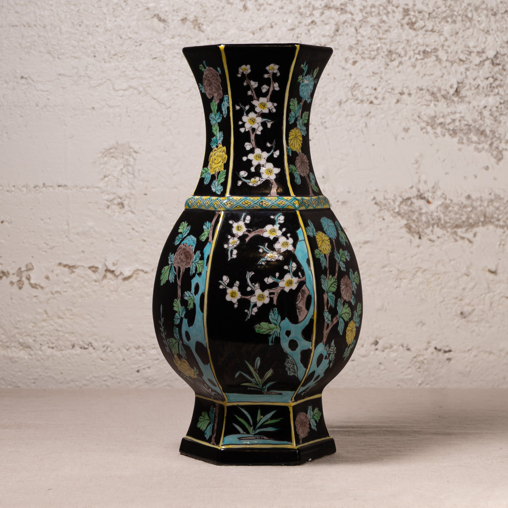 A black ceramic vase with a flared neck and hexagonal base, decorated with hand-painted floral motifs in turquoise, gold, green, and white, set against a textured off-white wall.