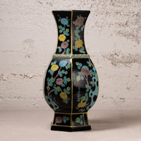 A black ceramic vase with a flared neck and hexagonal base, decorated with colorful floral motifs in yellow, blue, green, and brown, set against a textured off-white wall.