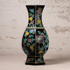 A black ceramic vase with a flared neck and hexagonal base, decorated with colorful floral motifs in yellow, blue, green, and brown, set against a textured off-white wall.