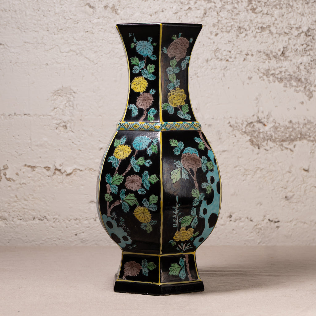 A black ceramic vase with a flared neck and hexagonal base, decorated with colorful floral motifs in yellow, blue, green, and brown, set against a textured off-white wall.