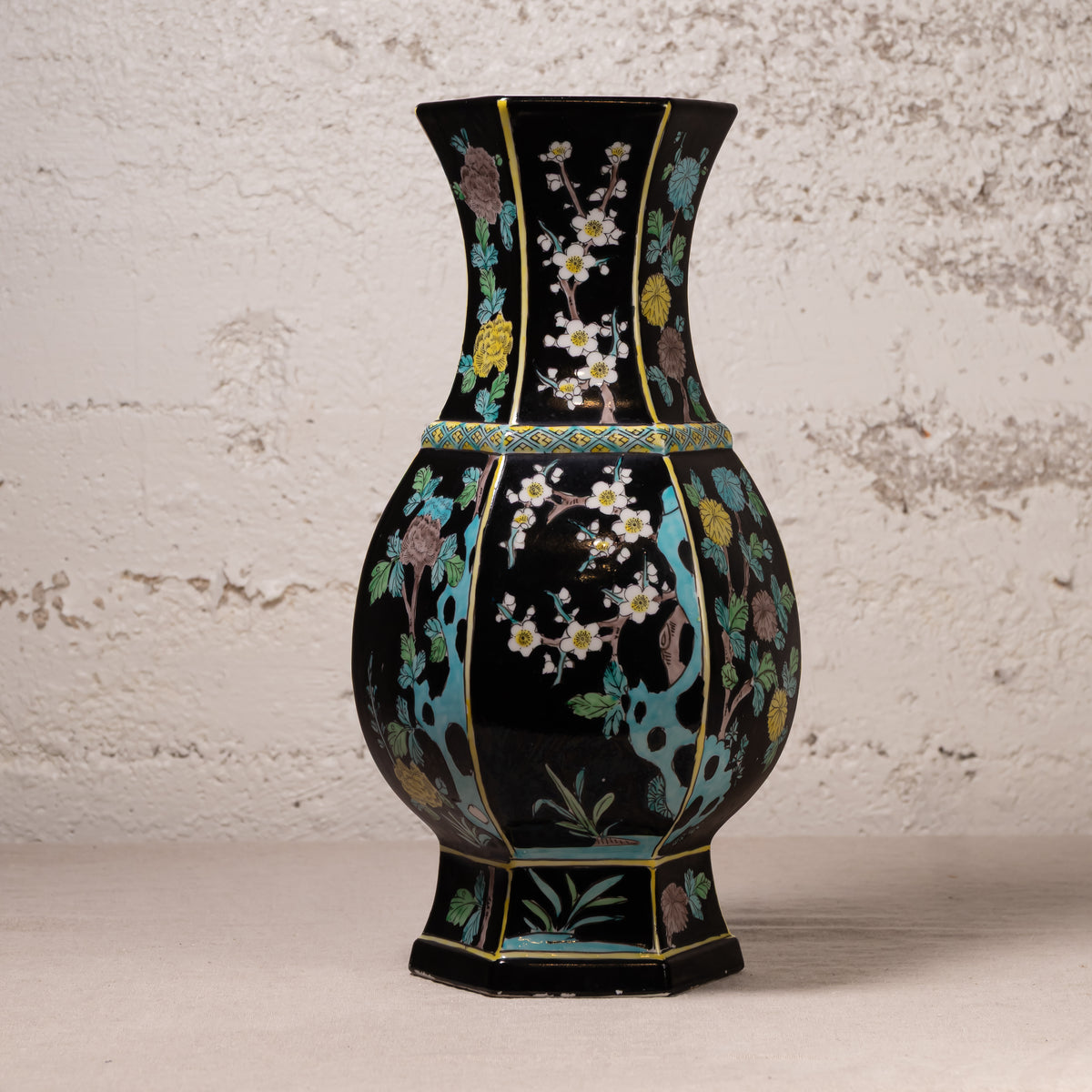 A black ceramic vase with hand-painted floral motifs in turquoise, gold, white, and pink, set against a textured off-white wall.