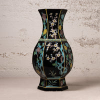 A black ceramic vase with hand-painted floral motifs in turquoise, gold, white, and pink, set against a textured off-white wall.