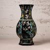 A black ceramic vase with hand-painted floral motifs in turquoise, gold, white, and pink, set against a textured off-white wall.