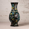 A black octagonal ceramic vase decorated with colorful floral motifs in yellow, green, and blue, set against a textured light beige wall.