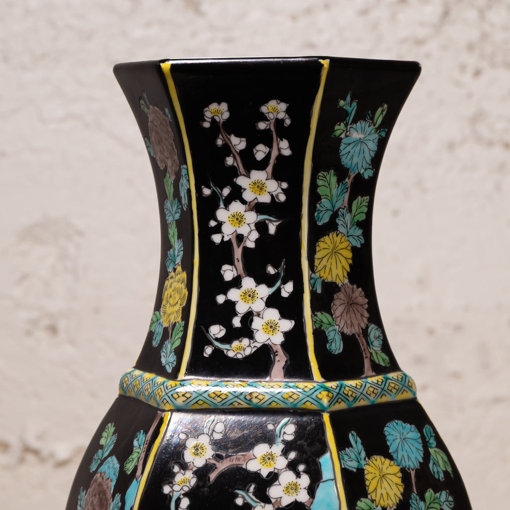 A close-up of a black ceramic vase decorated with hand-painted floral motifs in white, yellow, blue, and brown, featuring gold trim and a patterned band around the neck.