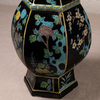 A close-up of a decorative ceramic vase with a glossy black glaze, featuring hand-painted floral motifs in turquoise, green, white, yellow, and pink, outlined with gold trim.