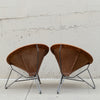 Two vintage-style leather armchairs with curved brown leather seats and thin metal legs, photographed side by side against a light concrete wall.