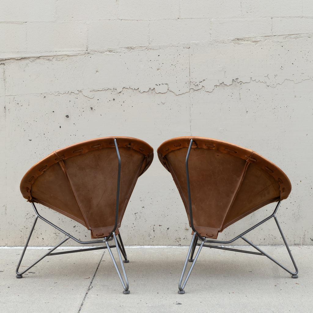 Two vintage-style leather armchairs with curved brown leather seats and thin metal legs, photographed side by side against a light concrete wall.