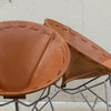 Two mid-century modern chairs with curved tan leather seats and silver metal wire frames, photographed against a light concrete wall.