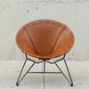 A mid-century modern accent chair with a warm brown leather seat and a black metal wire frame, set against a concrete wall.