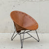 A mid-century modern leather chair with a warm tan-brown leather seat and a black metal tripod base, set against a textured concrete wall.