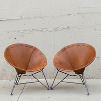 Two mid-century modern butterfly chairs with tan leather seats and gray metal legs, positioned side-by-side against a textured concrete wall.