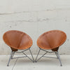 Two mid-century modern butterfly chairs with tan leather seats and gray metal legs, positioned side-by-side against a textured concrete wall.