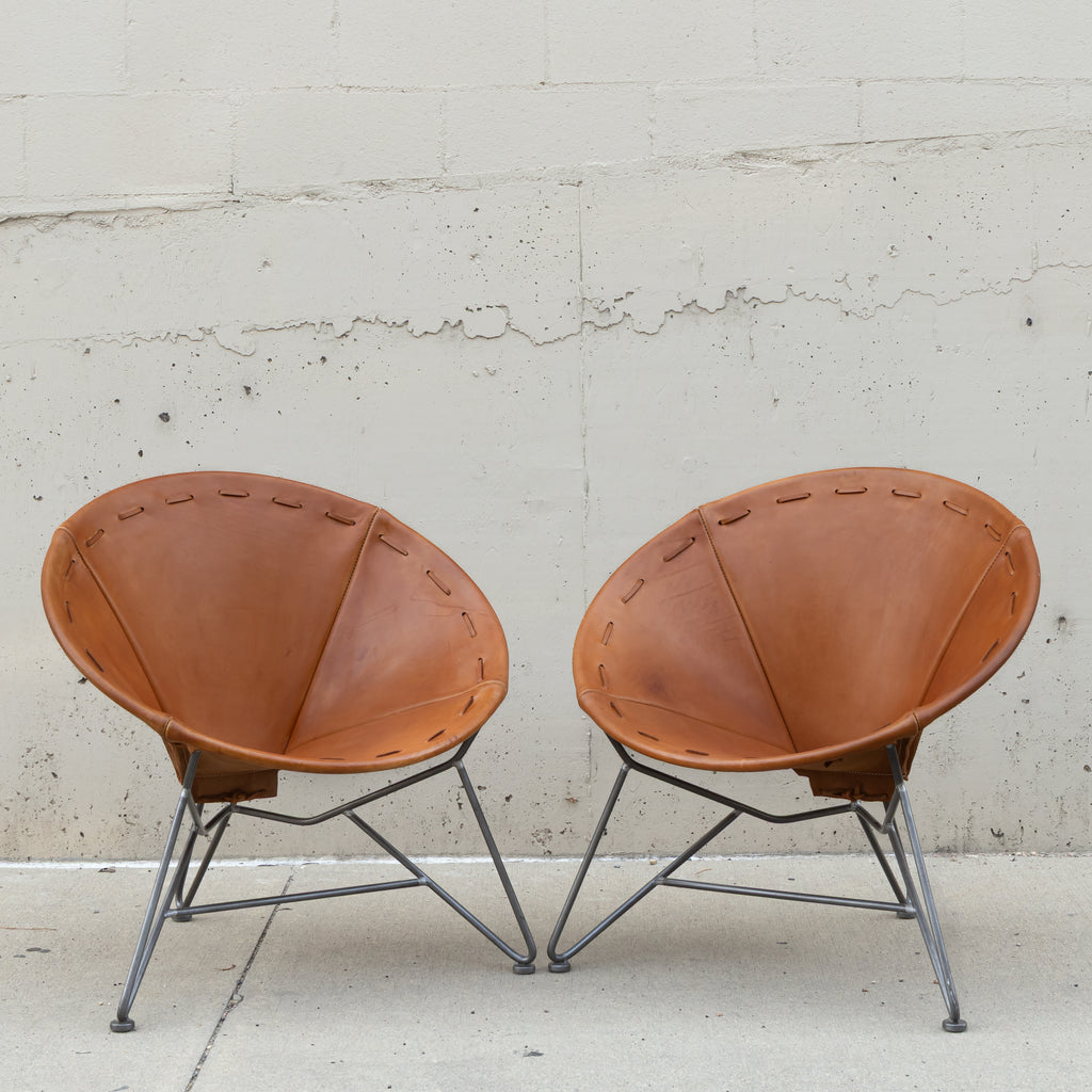 Two mid-century modern butterfly chairs with tan leather seats and gray metal legs, positioned side-by-side against a textured concrete wall.