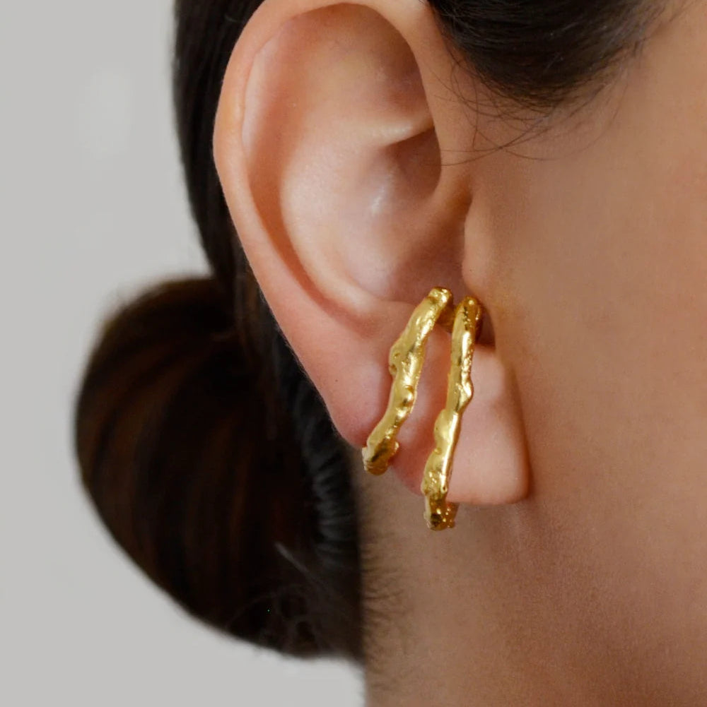 A close-up of a model's ear wearing a pair of textured, wavy gold hoop earrings.