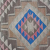 A close-up of a woven textile with a geometric Southwestern-style pattern, featuring a central diamond motif in blue, pink, and gray on a beige background, surrounded by zigzag bands of brown, gray, and pink.