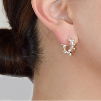 A close-up of a person's ear wearing a small silver huggie hoop earring with delicate dangling leaf-like details.