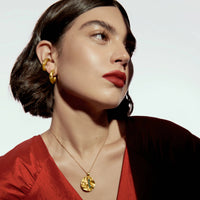 A close-up lifestyle shot of a model wearing a textured gold pendant necklace and matching gold hoop earrings, paired with a deep red satin V-neck top against a clean white background.
