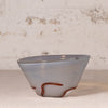 A handmade glass bowl with a light gray and white marbled pattern and a decorative reddish-brown swirl near the base, set against a rustic, textured off-white wall.