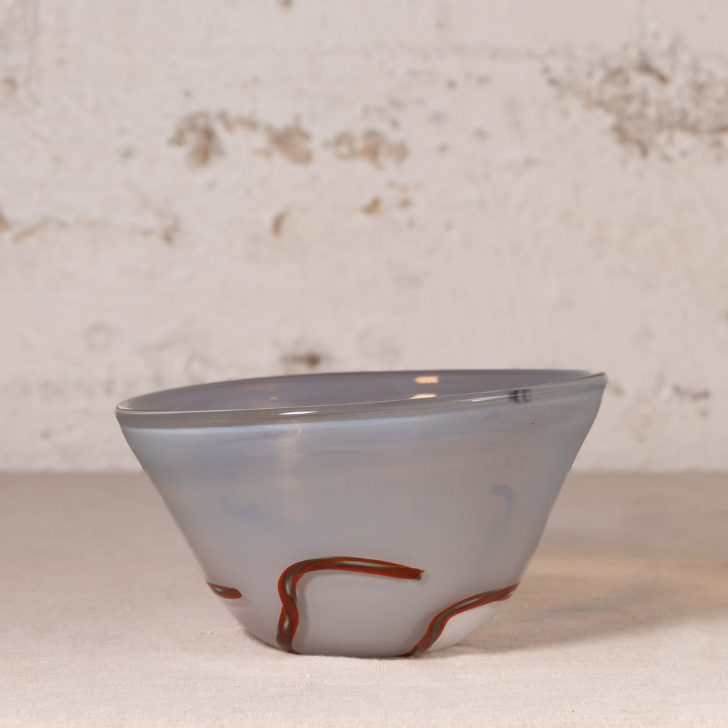 A handmade glass bowl with a light gray and white marbled pattern and a decorative reddish-brown swirl near the base, set against a rustic, textured off-white wall.