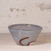 A handmade glass bowl with a frosted, semi-translucent pale blue-gray body and a thin, wavy reddish-brown glass accent near the base, set against a textured off-white background.
