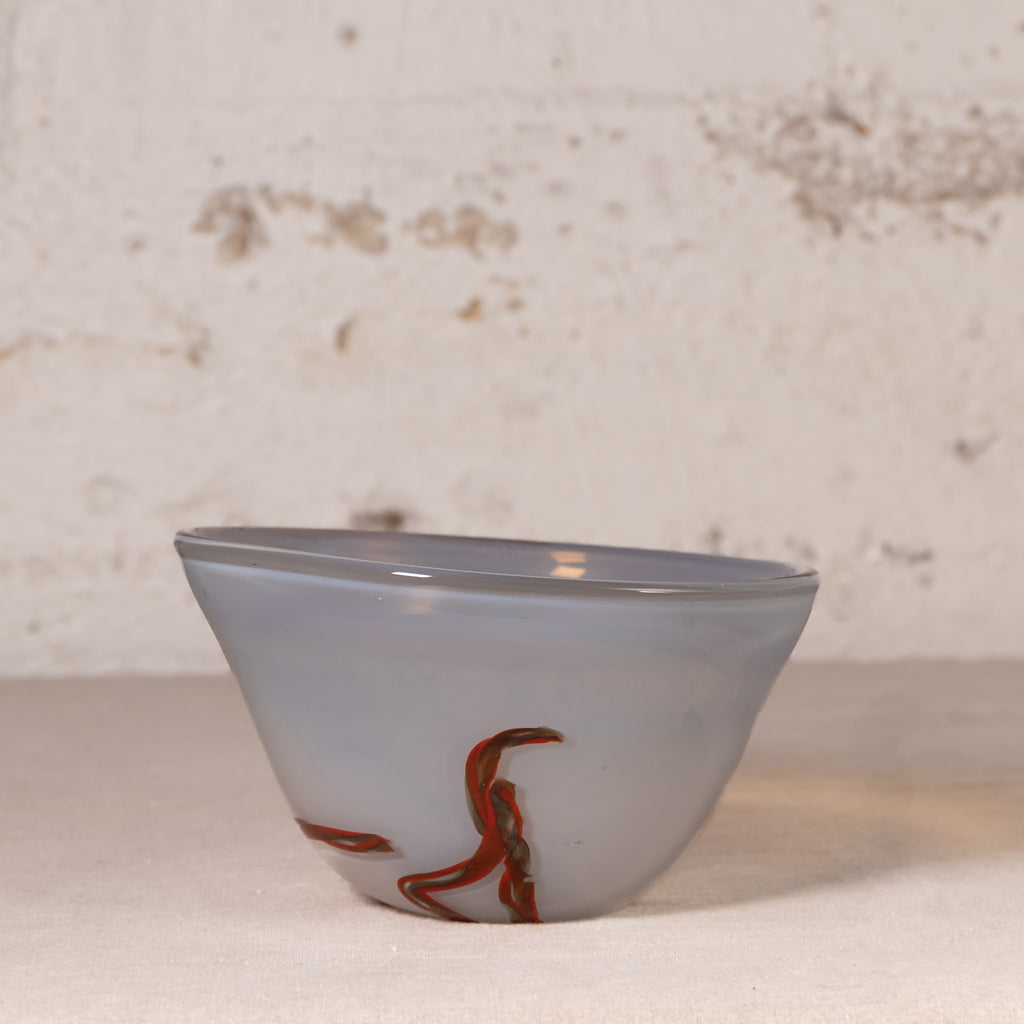 A small translucent glass bowl with a pale blue-gray body and red-orange abstract swirls near the base, photographed against a textured off-white wall and light surface.