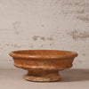 A rustic terracotta clay pedestal bowl with a textured, earthy orange-brown finish, set against a distressed off-white plaster wall.