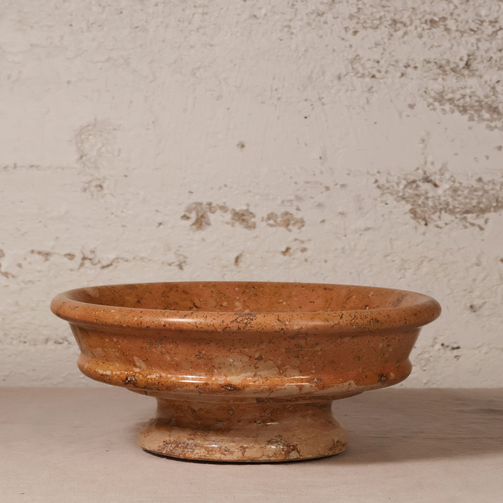 A rustic terracotta clay pedestal bowl with a textured, earthy orange-brown finish, set against a distressed off-white plaster wall.