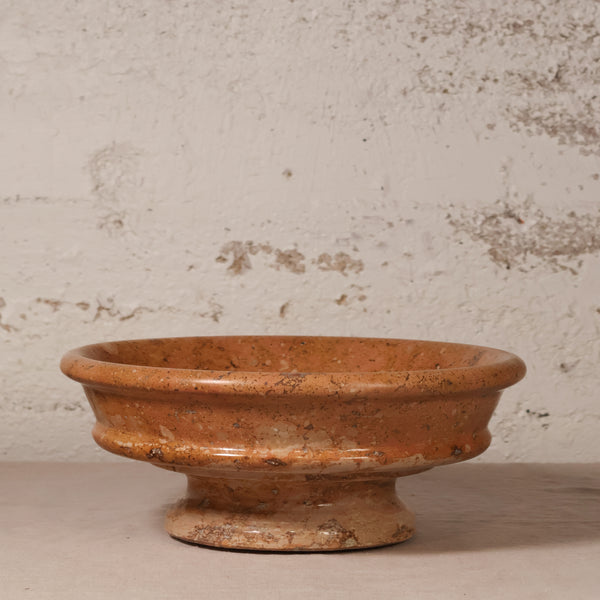 A rustic terracotta clay pedestal bowl with a textured, earthy orange-brown finish, set against a distressed off-white plaster wall.