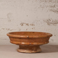 A rustic terracotta clay pedestal bowl with a textured, earthy orange-brown glaze, set against a distressed off-white plaster wall.