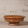 A rustic terracotta clay pedestal bowl with a textured, earthy orange-brown glaze, set against a distressed off-white plaster wall.